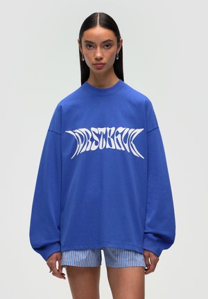 Blue oversized sweatshirt with wide sleeves and a ribbed crew neck, featuring bold white graphic text across the chest. Worn with striped shorts.