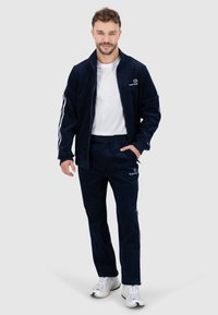 Navyblauw geribbeld trainingspak met witte strepen op de mouwen en broek. Het jack heeft een hoge kraag en een logo op de borst; gecombineerd met een wit t-shirt en sneakers.