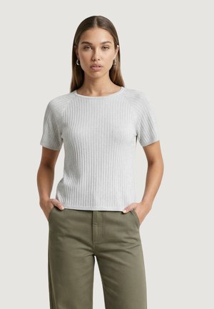 Donna con capelli lunghi e lisci che indossa orecchini a cerchio argentati, un top a maniche corte in maglia a coste grigio chiaro e pantaloni verde oliva con le mani nelle tasche.