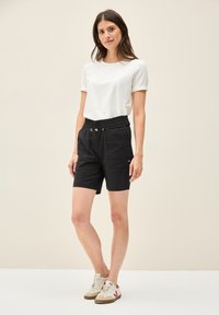 T-shirt bianca in cotone con maniche arrotolate, abbinata a pantaloni corti neri con vita elastica e tasche frontali. Il modello indossa scarpe da ginnastica beige e rosse.