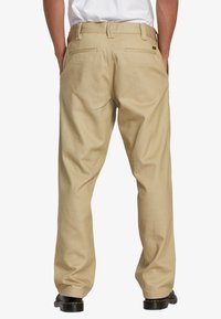 Pantalones beige de algodón, con un diseño de pierna recta, dos bolsillos traseros y una cinturilla estándar. Detalle de herrajes negros visible.