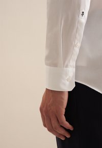 Witte longsleeve shirt met een gladde textuur, knoopmanchetten en een kleine zwart geborduurde bloem op de mouw.