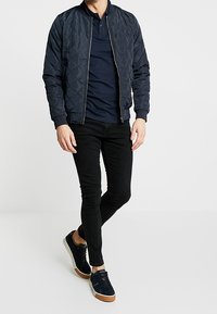 Laivastonsininen tikkikuvioitu bomber-takki vetoketjulla, mustat slim fit -farkut ja tummat lenkkitossut vaaleilla pohjilla. Teksturoitu kangas ja ribattu hihansuu.