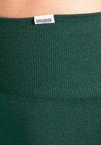 Grüner, gerippter Bund eines Kleidungsstücks mit einem weißen Etikett, auf dem "SMILODOX" steht. Die Stoffstruktur ist glatt und elastisch mit einheitlicher Farbe.