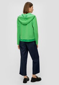 Zielona krótka bluza z kapturem, z krawatem, w połączeniu z luźnymi, ciemnymi dżinsami i czarnymi butami, widoczna z tyłu. Gładka faktura materiału.