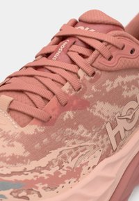 Rosa atletiska skor med textilstruktur, intrikat mönster över ovandelen och framträdande logotyp. Har robusta snören och dämpad sula.