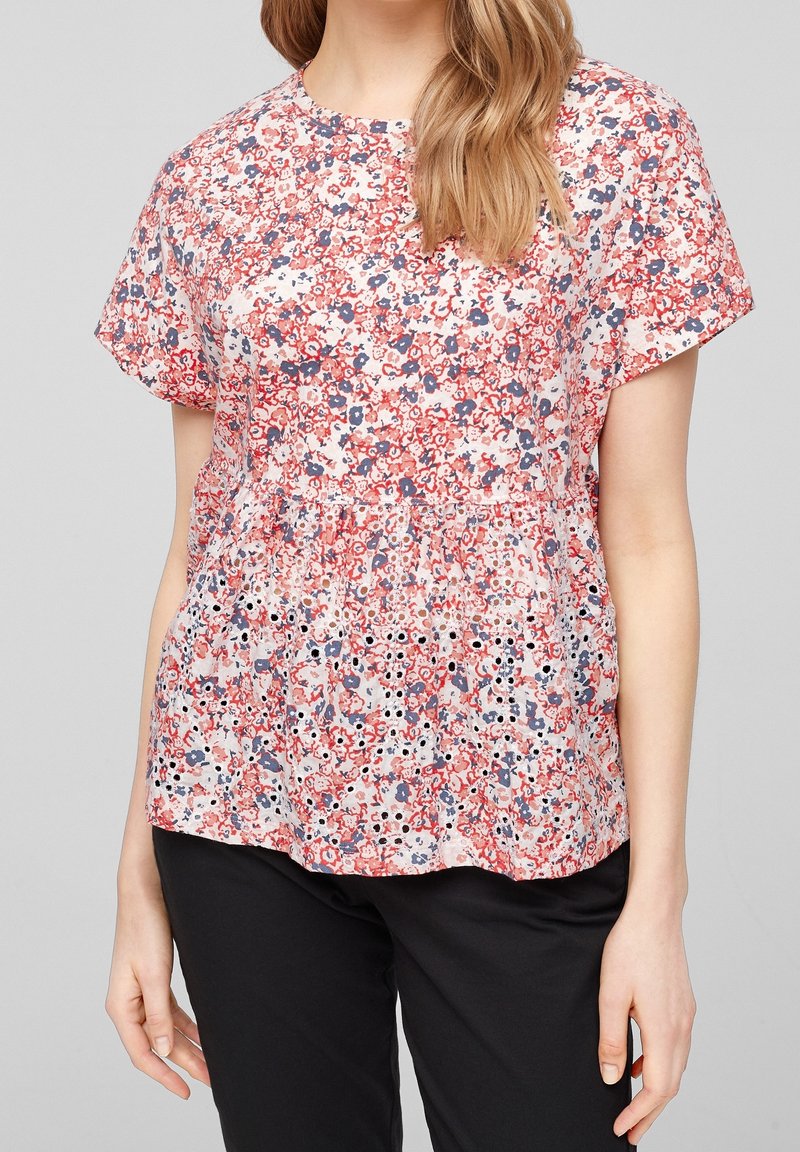 Blouse à motif floral aux teintes roses, rouges et bleues, confectionnée en tissu léger, dotée de manches courtes et d'un ourlet basque avec des détails en œillets.