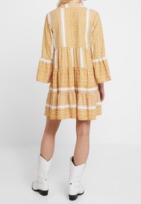 Gelbes gemustertes Kleid mit langen Glockenärmeln, gestuftem Design und weißen Akzenten; getragen mit weißen Stiefeletten im Western-Stil.