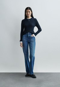 Γυναίκα που στέκεται με το ένα χέρι στην τσέπη, φοράει μπλε πουλόβερ με ριμπ υφή, λευκό υποκάμισο, μπλε τζιν ψηλόμεσα και μαύρα παπούτσια, μπροστά από γκρι φόντο.