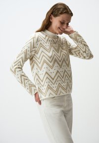 Crème trui met een zigzag- en stippenpatroon in goudtinten; gemaakt van zacht materiaal, met een hoge hals en lange mouwen. Gecombineerd met witte broeken.
