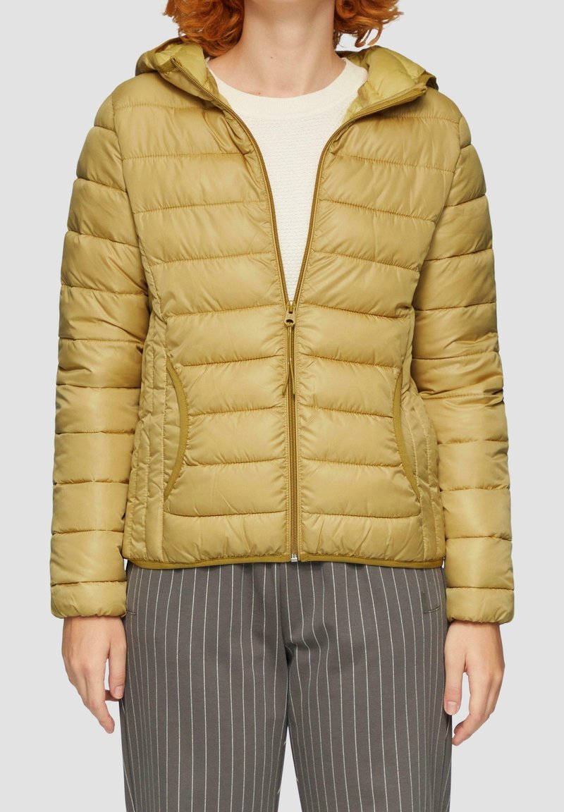 Personne portant une veste matelassée marron clair zippée avec capuche, un haut blanc en dessous et un pantalon gris à fines rayures.