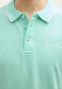 Mintgrön polo t-shirt med klassisk krage, tre knappar och vita accentlinjer. Texten "Tom Tailor" är broderad i vitt.