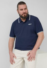 Marineblaues Poloshirt mit weißen Kragen- und Ärmelausschnitten, aus Baumwolle gefertigt. Mit einem Logo auf der linken Brustseite und einem klassischen Schnitt.