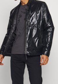Giacca leggera nera tipo puffer con finitura lucida, colletto a coste, tasche frontali con zip e una silhouette moderna e aderente.
