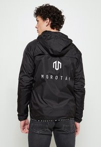 Veste légère noire avec une capuche, présentant un logo blanc et le texte "MOROTAI" au dos. Le tissu a une texture lisse et brillante.