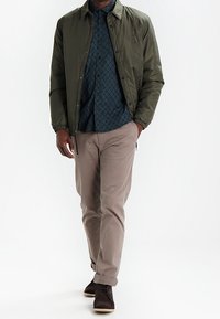 Olijfgroene lichte bomberjack, gestreept donkerblauw shirt, beige broek en bruine schoenen. Jack heeft elastische manchetten en een logo-detail.