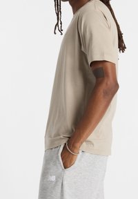 Beige bavlněné tričko s krátkými rukávy, kulatým výstřihem a volným střihem. Disponuje hladkým povrchem a minimálním švem, nošeno s šedými tepláky.