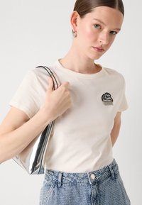Cream short-sleeve t-shirt with a black and silver embroidered design, paired with light blue denim jeans and a shiny silver handbag.
