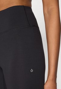 Svarte leggings med høyt liv laget av glatt, elastisk stoff. Har flatlocksøm og en subtil logo på venstre side.