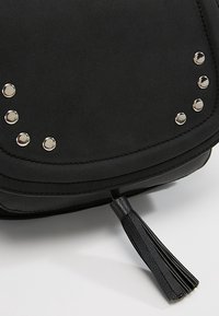 Schwarze Tasche mit einer weichen, strukturierten Oberfläche, die mit silbernen Nieten und einem dekorativen Quast am Reißverschluss verziert ist. Sauberes und modernes Design.