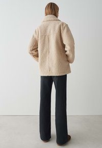Veste en fausse fourrure beige et fluffy avec un col, des détails de couture au dos et une coupe décontractée, associée à un pantalon évasé noir et des chaussures marron.