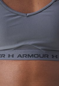 Sostén deportivo gris con un tejido suave, un escote redondeado y una banda elástica negra con logo en la parte inferior. Detalles texturizados en el material.