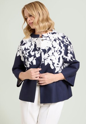 Mujer con cabello rubio hasta los hombros abotonándose una chaqueta azul marino con patrón de flores blancas, vistiendo pantalones blancos frente a un fondo claro.