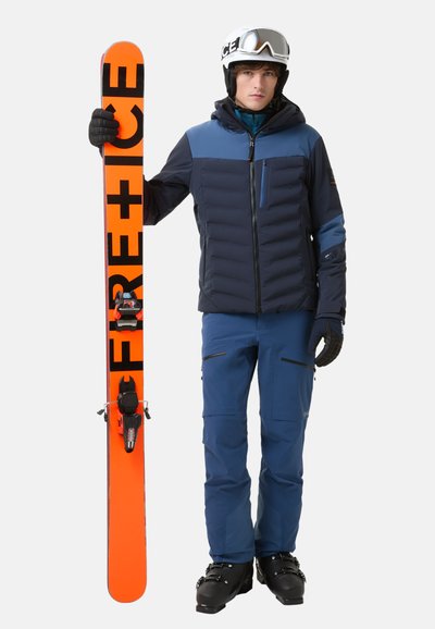 Lyžařský outfit zahrnující tmavě modrou nadýchanou bundu, odpovídající modré kalhoty, černé rukavice a bílou helmu. Drží oranžové lyže s čern�ým nápisem.