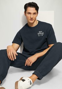 Marineblauw t-shirt met korte mouwen met de opdruk "24/7 Planking Club" en bijpassende sweatpants. Witte sneakers met bruine accenten maken de outfit compleet.