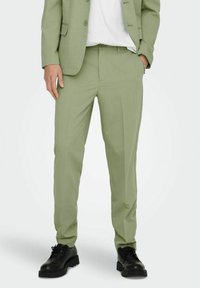 Pantalones verde claro hechos de una tela suave, con corte entallado, con pliegues frontales, bolsillos laterales y combinados con zapatos negros de cordones.