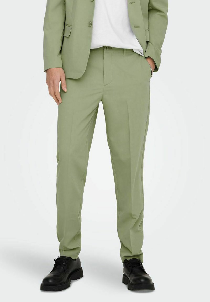 Pantalones verde claro hechos de una tela suave, con corte entallado, con pliegues frontales, bolsillos laterales y combinados con zapatos negros de cordones.