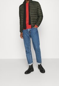 Groene pufferjack, rode sweater, donkere sjaal, blauwe jeans, opgerolde mouwen, zwarte enkelboots. Zachte texturen met doorstikte details.