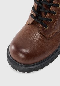 Boot din piele maro texturată, cu șireturi groase negre, ochiuri din alamă și o talpă robustă din cauciuc negru.