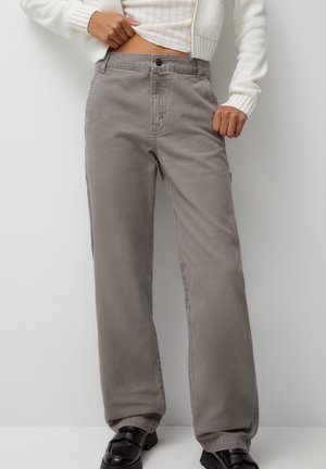 Persoon draagt grijze broek met wijde pijpen, zwarte schoenen en een witte trui, en tilt de trui licht op om het taillegebied te laten zien.