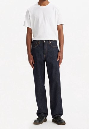 Homme portant un t-shirt blanc uni à manches courtes, un jean droit bleu foncé et des chaussures en cuir noires, debout devant un fond blanc.
