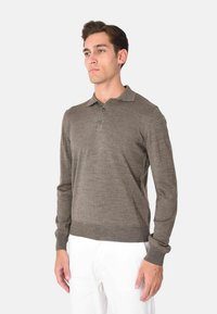 Polo a maniche lunghe marrone realizzato in tessuto morbido; presenta un colletto classico, una chiusura con tre bottoni e polsini a coste. Indossato con pantaloni bianchi.