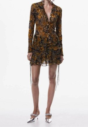 Model wearing a fitted long-sleeve button-up blouse and matching short skirt with brown and black floral pattern, paired with silver pointed heels.