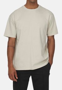 Helles Beige, Baumwoll-T-Shirt mit kurzen Ärmeln, Rundhalsausschnitt, lässigem Schnitt und glatter Textur. Getragen mit schwarzen Hosen.