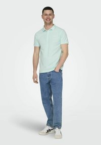 Hellgrünes Polohemd, kurzärmlig, mit dezentem Logo. Getragen mit hellblauen Jeans und weißen Sneakers mit schwarzen Akzenten.