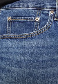 Wąskie niebieskie dżinsy z denimu, pokazujące zbliżenie na przednią kieszeń z złotym przeszyciem i metalowymi nitami, z efektem wyblakłej tekstury.