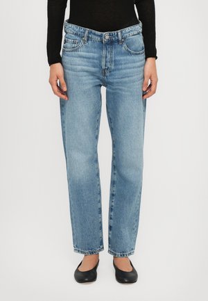 Personne portant un jean bleu clair taille haute à jambes droites, un haut noir à manches longues et des chaussures plates noires, debout devant un fond uni.