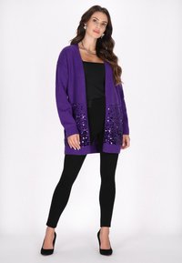 Cardigan lavorato a maglia viola con tasche decorate con paillettes, abbinato a leggings neri e tacchi alti. Design semplice con apertura frontale.