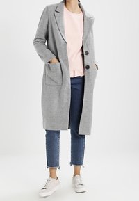 Cappotto lungo grigio realizzato in un tessuto morbido, con colletto a risvolto, due tasche frontali e bottoni neri. Indossato sopra una camicia rosa chiaro e jeans blu.