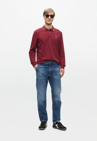 Rode polo shirt met witte kraagdetails, gecombineerd met blauwe spijkerbroeken en zwarte sneakers. Model draagt een zonnebril en staat rechtop.