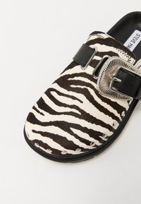 Black and white zebra print clog featuring a black strap and a silver buckle, with studded accents around the edge and a textured surface.
