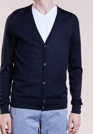 Homme portant un cardigan boutonné bleu marine foncé par-dessus un t-shirt blanc à col en V et un pantalon gris clair, debout devant un fond uni.