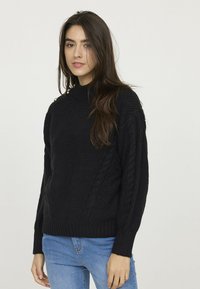 Pull en tricot noir avec un col montant, doté de détails en mailles torsadées sur les manches et d'un ourlet côtelé. Associé à un jean bleu.