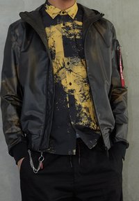 Person som bär en svart bomberjacka över en svart och gul mönstrad skjorta med knappar, med händerna i fickorna på svarta byxor mot en grå vägg.