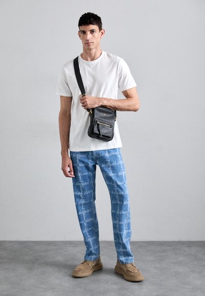 Bílý bavlněný tričko, modré džínové kalhoty s bílým nápisem, černá kožená kabelka přes rameno, hnědé semišové mokasíny, šedé pozadí.