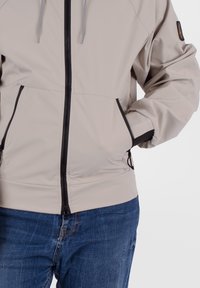 Giacca zip-up beige chiaro con una texture liscia, zip nera e cordini regolabili. Presenta tasche laterali e polsini elasticizzati.
